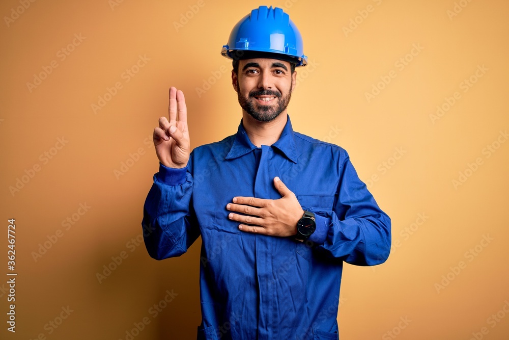 Mechanic man with beard wearing blue uniform and safety helmet over yellow background smiling swearing with hand on chest and fingers up, making a loyalty promise oath