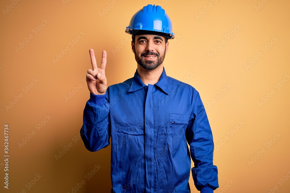 Mechanic man with beard wearing blue uniform and safety helmet over yellow background showing and pointing up with fingers number two while smiling confident and happy.