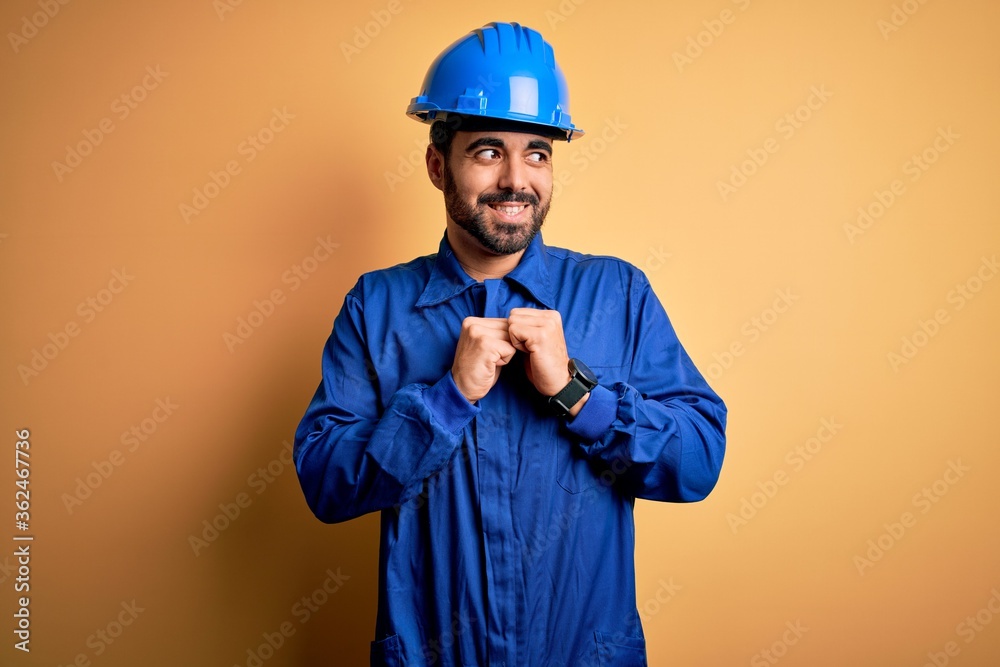 Mechanic man with beard wearing blue uniform and safety helmet over yellow background laughing nervous and excited with hands on chin looking to the side