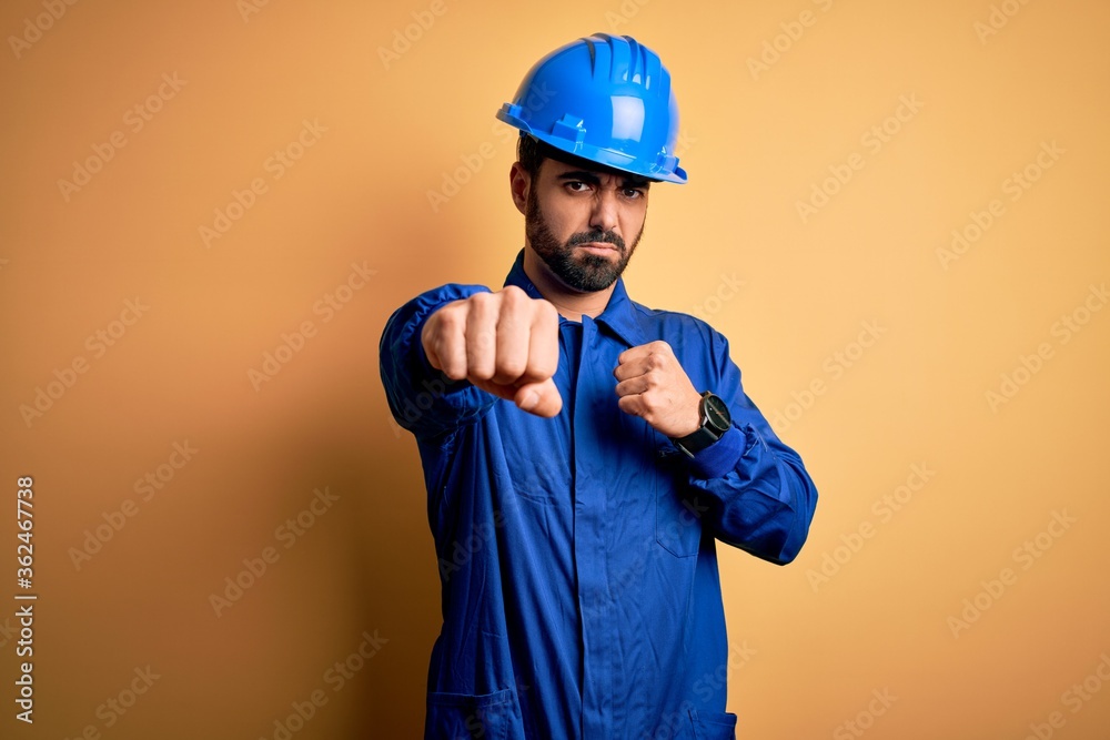 Mechanic man with beard wearing blue uniform and safety helmet over yellow background Punching fist to fight, aggressive and angry attack, threat and violence