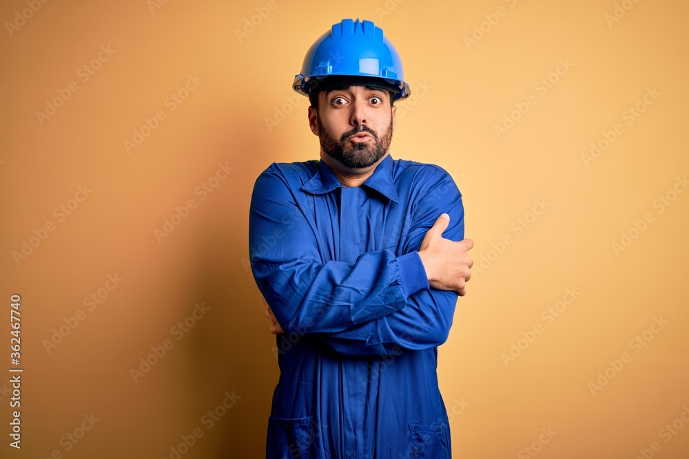 Mechanic man with beard wearing blue uniform and safety helmet over yellow background shaking and freezing for winter cold with sad and shock expression on face