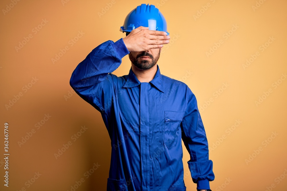 Mechanic man with beard wearing blue uniform and safety helmet over yellow background covering eyes with hand, looking serious and sad. Sightless, hiding and rejection concept
