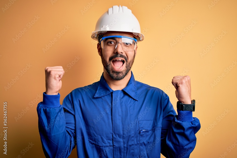 Mechanic man with beard wearing blue uniform and safety glasses over yellow background celebrating surprised and amazed for success with arms raised and open eyes. Winner concept.