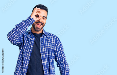 Young hispanic man wearing casual clothes doing ok gesture with hand smiling, eye looking through fingers with happy face.