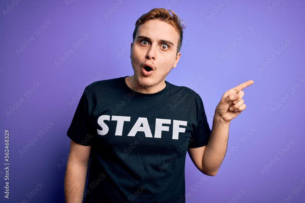 Young handsome redhead worker man wearing staff t-shirt uniform over purple background Surprised pointing with finger to the side, open mouth amazed expression.