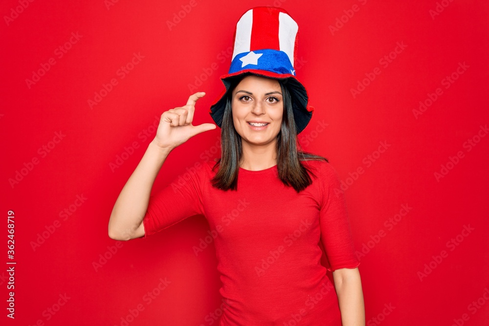 Young beautiful brunette woman wearing united states hat celebrating independence day smiling and confident gesturing with hand doing small size sign with fingers looking and the camera. Measure. Young beautiful brunette woman wearing united states hat celebrating independence day smiling and confident gesturing with hand doing small size sign with fingers looking and the camera. Measure.