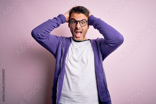 Young handsome man wearing purple sweatshirt and glasses standing over pink background Crazy and scared with hands on head, afraid and surprised of shock with open mouth