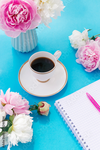 Summer romantic breakfast. Blue vase with peonies, white cup of coffee, notepad for entries with a pen on a blue background.Good morning concept.Copy space, selective focus with shallow depth of field