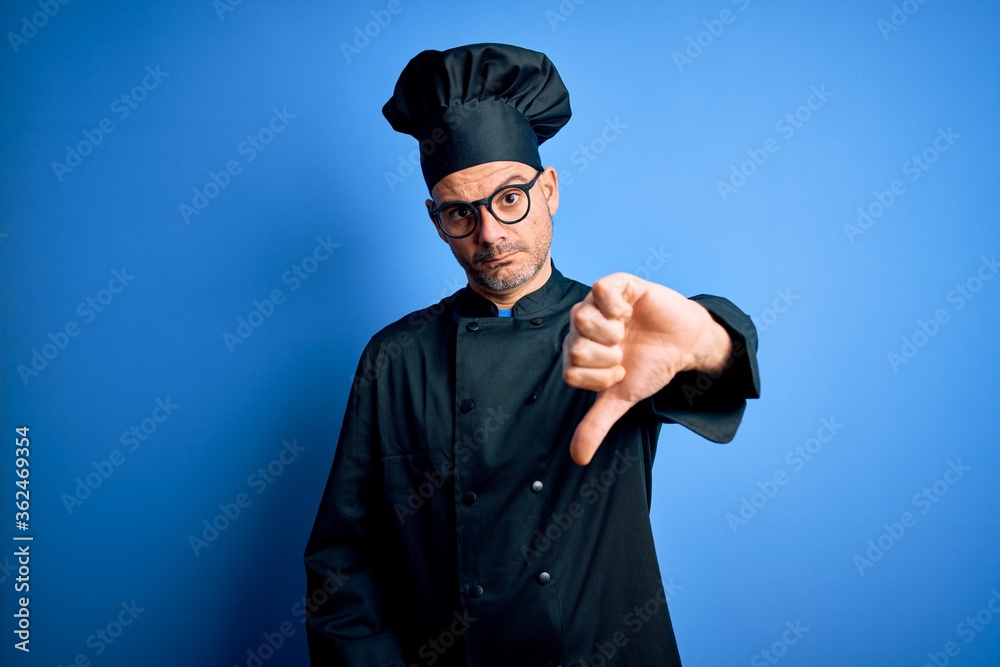 Young handsome chef man wearing cooker uniform and hat over isolated blue background looking unhappy and angry showing rejection and negative with thumbs down gesture. Bad expression. Young handsome chef man wearing cooker uniform and hat over isolated blue background looking unhappy and angry showing rejection and negative with thumbs down gesture. Bad expression.