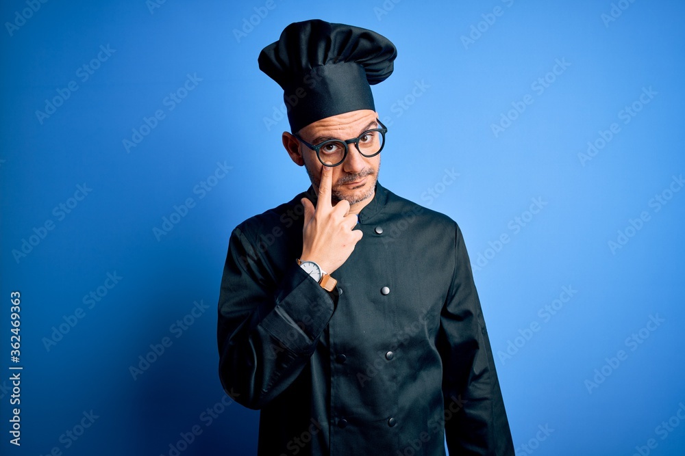 Young handsome chef man wearing cooker uniform and hat over isolated blue background Pointing to the eye watching you gesture, suspicious expression Young handsome chef man wearing cooker uniform and hat over isolated blue background Pointing to the eye watching you gesture, suspicious expression