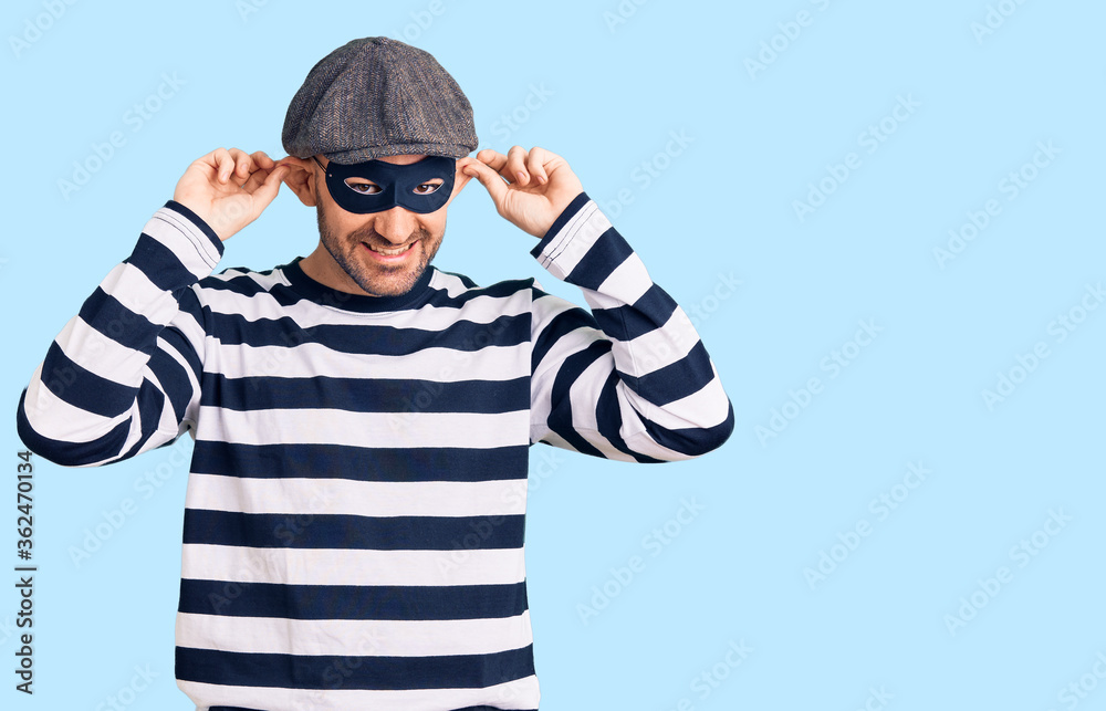 Young handsome man wearing burglar mask smiling pulling ears with fingers, funny gesture. audition problem Young handsome man wearing burglar mask smiling pulling ears with fingers, funny gesture. audition problem