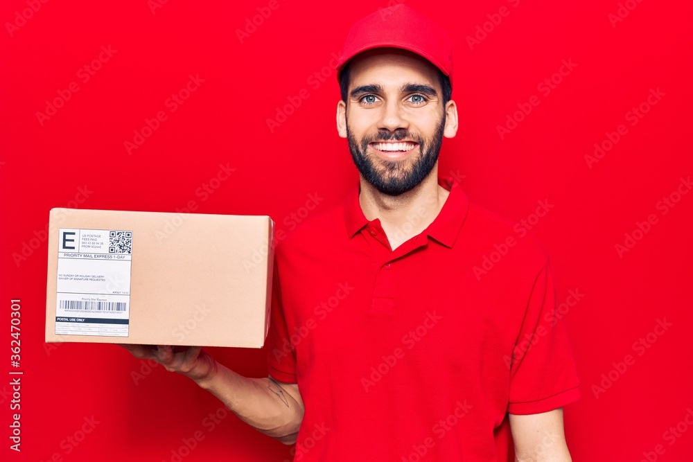 Young handsome man with beard wearing delivery uniform holding package looking positive and happy standing and smiling with a confident smile showing teeth Young handsome man with beard wearing delivery uniform holding package looking positive and happy standing and smiling with a confident smile showing teeth