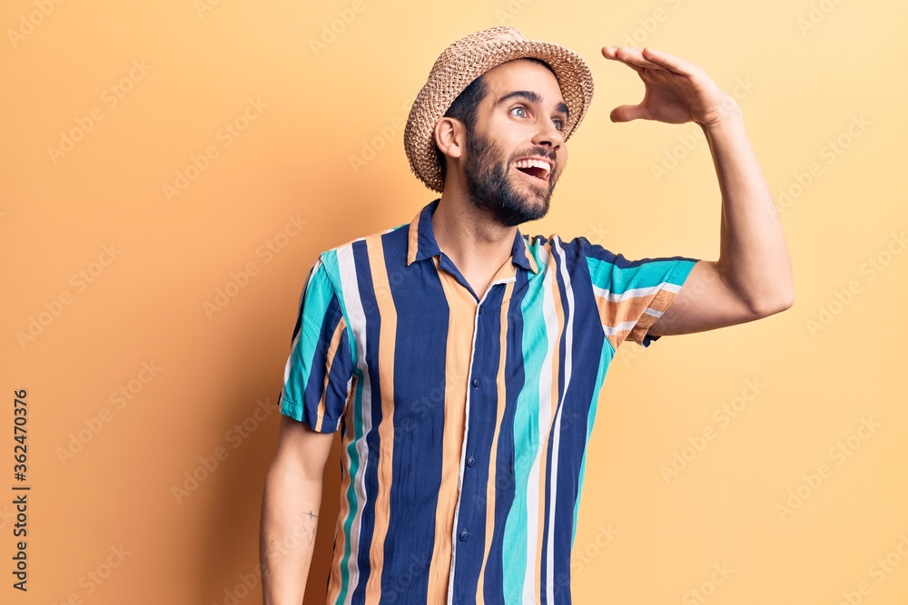 Young handsome man with beard wearing summer hat and shirt very happy and smiling looking far away with hand over head. searching concept. Young handsome man with beard wearing summer hat and shirt very happy and smiling looking far away with hand over head. searching concept.