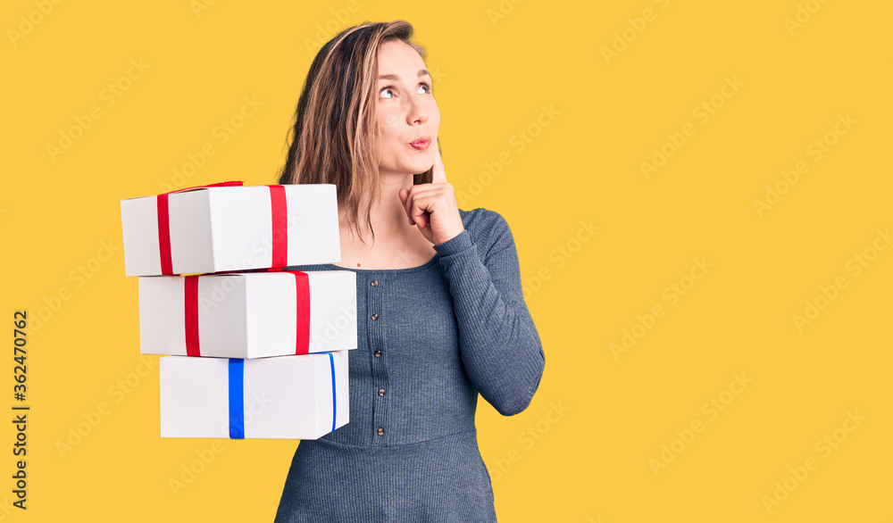 Young beautiful blonde woman holding birthday gift serious face thinking about question with hand on chin, thoughtful about confusing idea Young beautiful blonde woman holding birthday gift serious face thinking about question with hand on chin, thoughtful about confusing idea