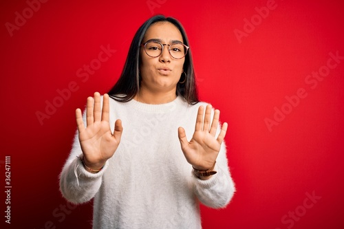 Young beautiful asian woman wearing casual sweater and glasses over red background Moving away hands palms showing refusal and denial with afraid and disgusting expression. Stop and forbidden. © Krakenimages.com