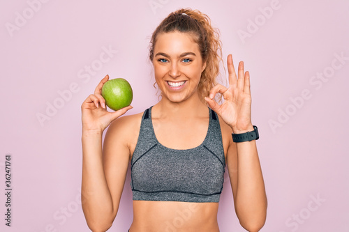 Young beautiful blonde sporty woman with blue eyes holding healthy green apple fruit doing ok sign with fingers, excellent symbol