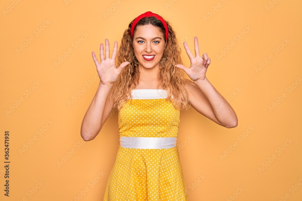 Beautiful blonde pin-up woman with blue eyes wearing diadem standing over yellow background showing and pointing up with fingers number eight while smiling confident and happy.