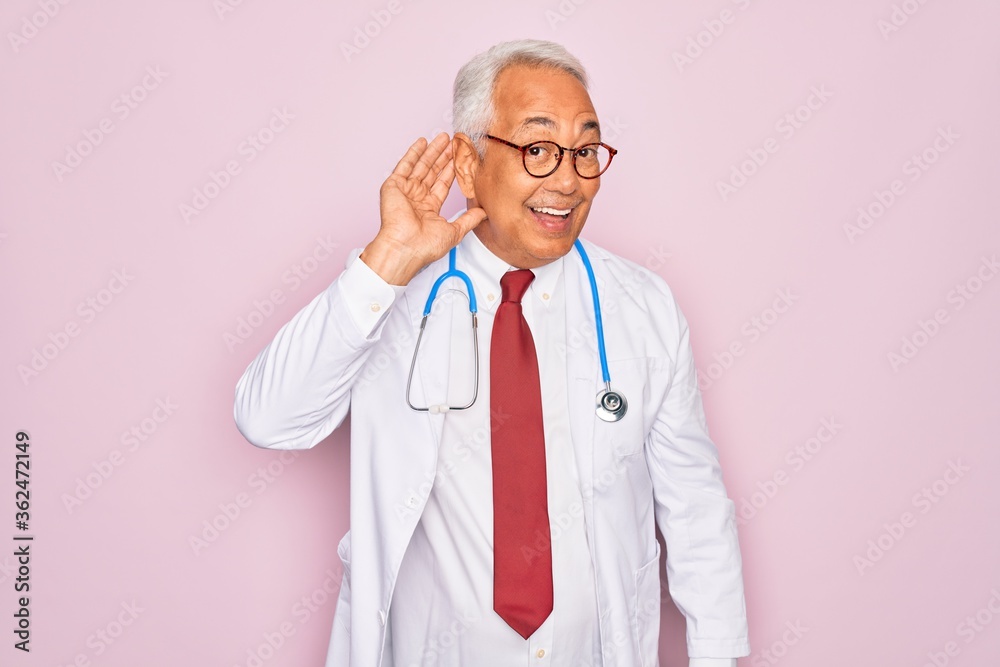 Middle age senior grey-haired doctor man wearing stethoscope and professional medical coat smiling with hand over ear listening an hearing to rumor or gossip. Deafness concept. Middle age senior grey-haired doctor man wearing stethoscope and professional medical coat smiling with hand over ear listening an hearing to rumor or gossip. Deafness concept.