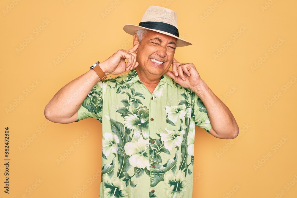 Middle age senior grey-haired man wearing summer hat and floral shirt on beach vacation covering ears with fingers with annoyed expression for the noise of loud music. Deaf concept. Middle age senior grey-haired man wearing summer hat and floral shirt on beach vacation covering ears with fingers with annoyed expression for the noise of loud music. Deaf concept.