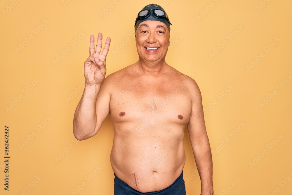 Middle age senior grey-haired swimmer man wearing swimsuit, cap and goggles showing and pointing up with fingers number three while smiling confident and happy.