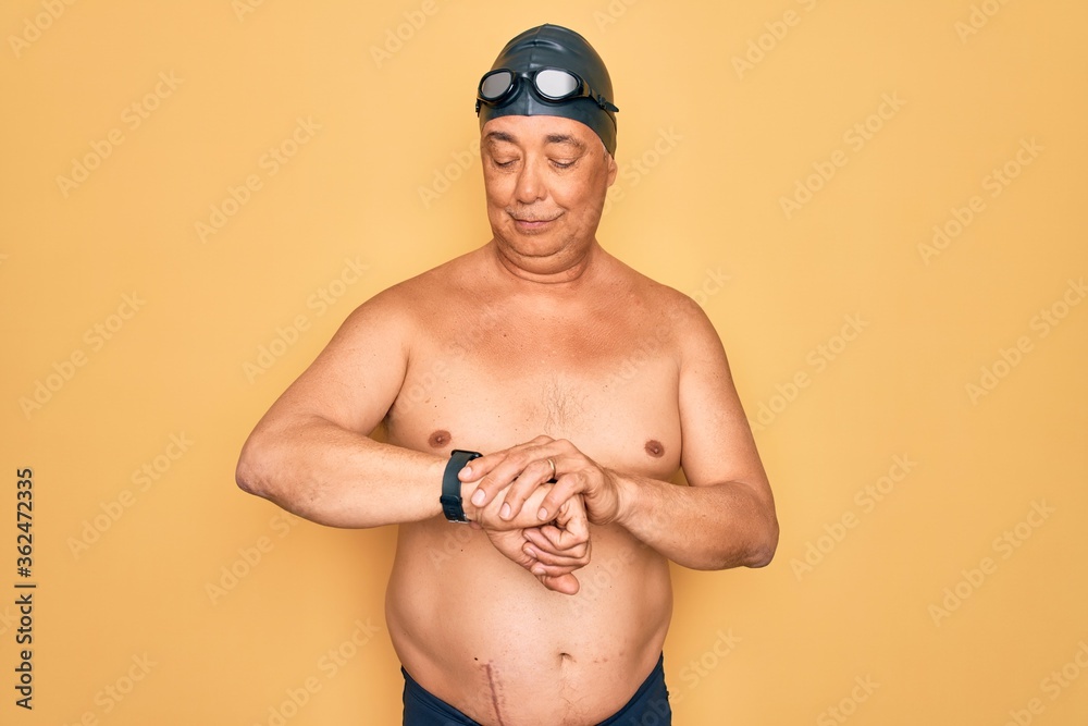Middle age senior grey-haired swimmer man wearing swimsuit, cap and goggles Checking the time on wrist watch, relaxed and confident