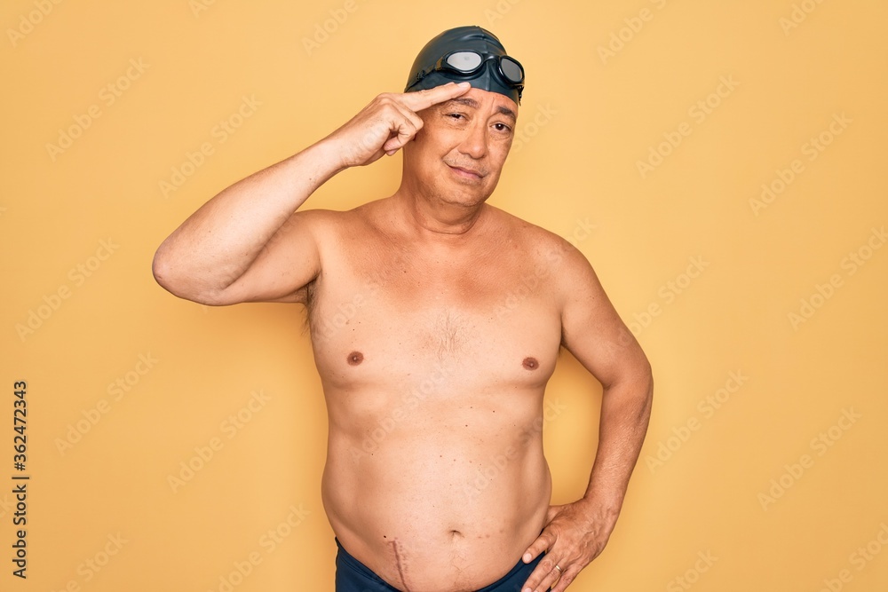 Middle age senior grey-haired swimmer man wearing swimsuit, cap and goggles pointing unhappy to pimple on forehead, ugly infection of blackhead. Acne and skin problem