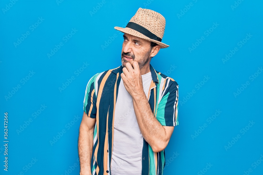 Middle age handsome tourist man on vacation wearing shirt and hat over blue background thinking concentrated about doubt with finger on chin and looking up wondering Middle age handsome tourist man on vacation wearing shirt and hat over blue background thinking concentrated about doubt with finger on chin and looking up wondering