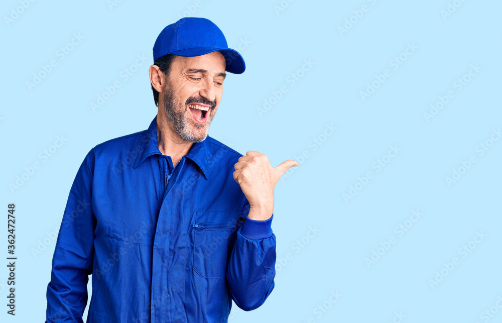 Middle age handsome man wearing mechanic uniform smiling with happy face looking and pointing to the side with thumb up.