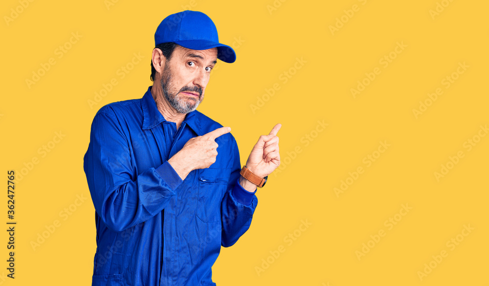 Middle age handsome man wearing mechanic uniform pointing aside worried and nervous with both hands, concerned and surprised expression