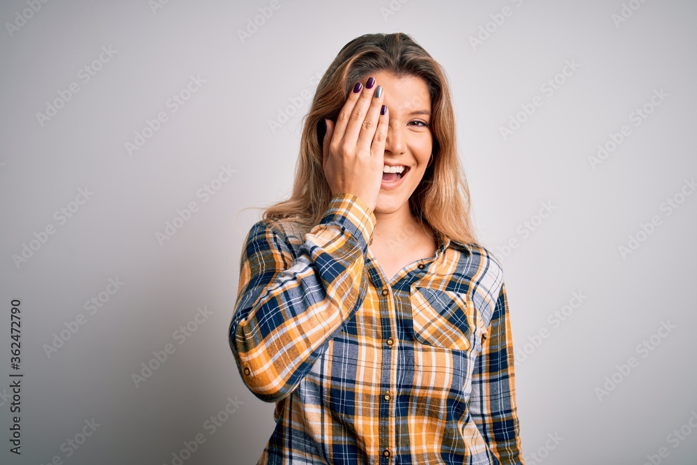 Young beautiful blonde woman wearing casual shirt standing over isolated white background covering one eye with hand, confident smile on face and surprise emotion. Young beautiful blonde woman wearing casual shirt standing over isolated white background covering one eye with hand, confident smile on face and surprise emotion.