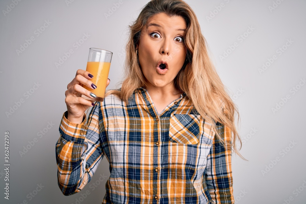 Young beautiful blonde woman drinking glass of healthy orange juice over white background scared in shock with a surprise face, afraid and excited with fear expression