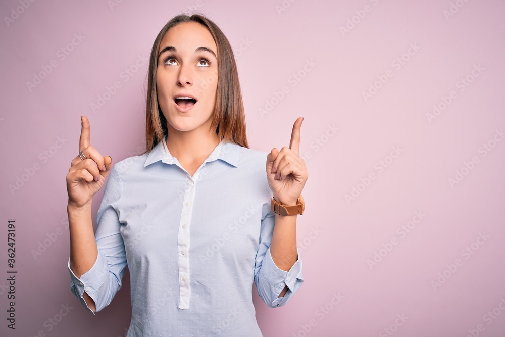 Young beautiful businesswoman wearing elegant shirt standing over isolated pink background amazed and surprised looking up and pointing with fingers and raised arms. Young beautiful businesswoman wearing elegant shirt standing over isolated pink background amazed and surprised looking up and pointing with fingers and raised arms.