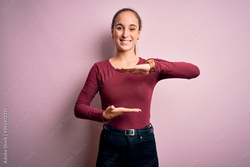 Young beautiful woman wearing casual t-shirt standing over isolated pink background gesturing with hands showing big and large size sign, measure symbol. Smiling looking at the camera. Measuring. Young beautiful woman wearing casual t-shirt standing over isolated pink background gesturing with hands showing big and large size sign, measure symbol. Smiling looking at the camera. Measuring.