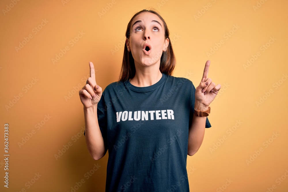 Young beautiful woman wearing volunteer t-shirt doing volunteering over yellow background amazed and surprised looking up and pointing with fingers and raised arms. Young beautiful woman wearing volunteer t-shirt doing volunteering over yellow background amazed and surprised looking up and pointing with fingers and raised arms.