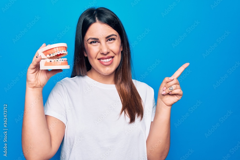 Young beautiful brunette woman asking for dental care holding denture with backets smiling happy pointing with hand and finger to the side Young beautiful brunette woman asking for dental care holding denture with backets smiling happy pointing with hand and finger to the side