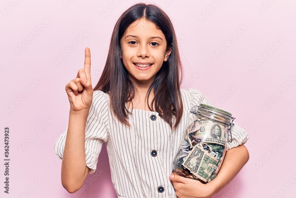 Beautiful child girl holding jar with savings smiling with an idea or question pointing finger with happy face, number one Beautiful child girl holding jar with savings smiling with an idea or question pointing finger with happy face, number one