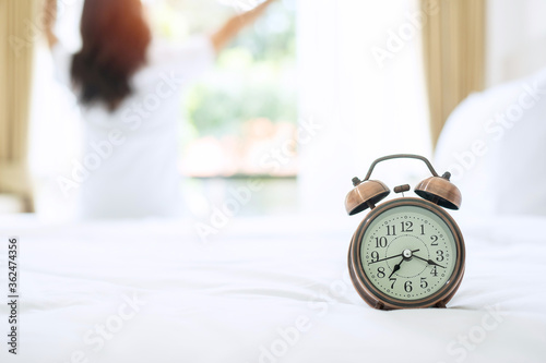 Closeup alarm clock with background of happy woman stretching in bed after waking up, young adult female rising arms and looking to window in the morning. fresh relax and have a nice day concepts