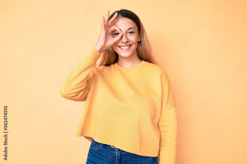 Young brunette woman over yellow background smiling happy doing ok sign with hand on eye looking through fingers Young brunette woman over yellow background smiling happy doing ok sign with hand on eye looking through fingers
