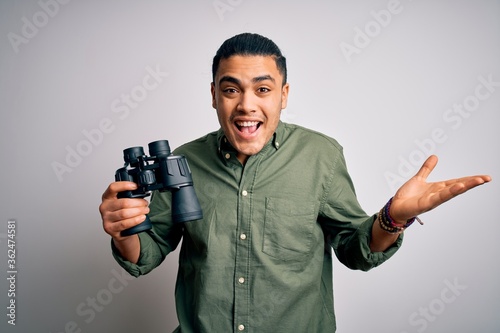 Young brazilian man looking through binoculars over isolated white background very happy and excited, winner expression celebrating victory screaming with big smile and raised hands