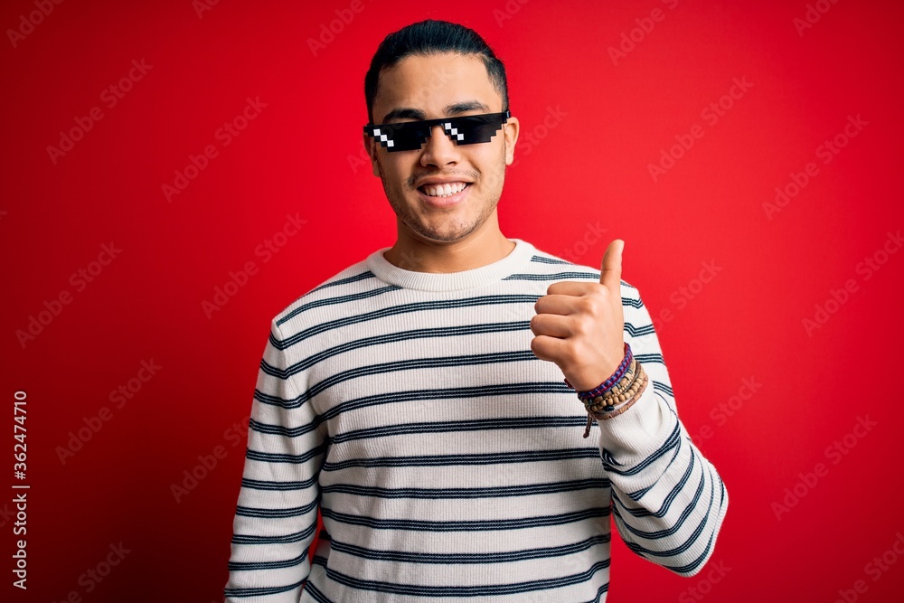 Φωτογραφία Young brazilian man wearing funny thug life sunglasses over isolated red background doing happy thumbs up gesture with hand