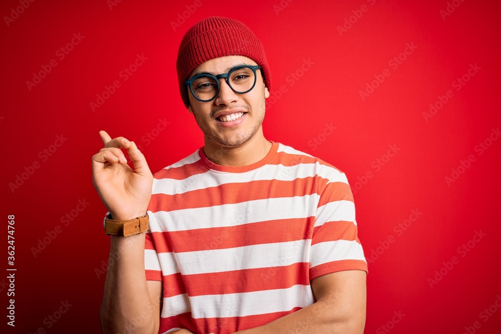 Φωτογραφία Young brazilian man wearing casual striped t-shirt and glasses over isolated red background with a big smile on face, pointing with hand and finger to the side looking at the camera