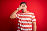 Young brazilian man wearing casual striped t-shirt standing over isolated red background peeking in shock covering face and eyes with hand, looking through fingers with embarrassed expression.