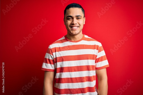 Young brazilian man wearing casual striped t-shirt standing over isolated red background with a happy and cool smile on face. Lucky person.