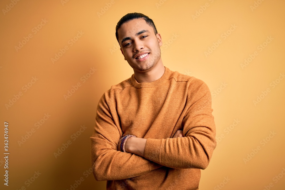 Young brazilian man wearing casual sweater standing over isolated yellow background happy face smiling with crossed arms looking at the camera. Positive person. Young brazilian man wearing casual sweater standing over isolated yellow background happy face smiling with crossed arms looking at the camera. Positive person.