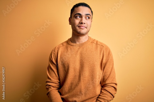 Young brazilian man wearing casual sweater standing over isolated yellow background smiling looking to the side and staring away thinking.