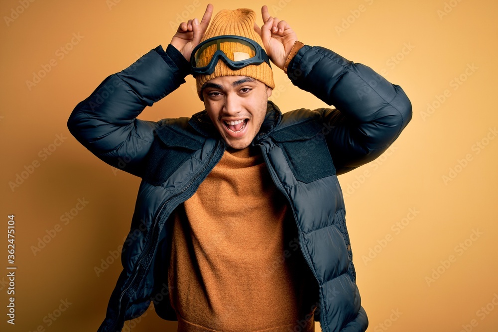 Young brazilian skier man wearing snow sportswear and ski goggles over yellow background Posing funny and crazy with fingers on head as bunny ears, smiling cheerful Young brazilian skier man wearing snow sportswear and ski goggles over yellow background Posing funny and crazy with fingers on head as bunny ears, smiling cheerful