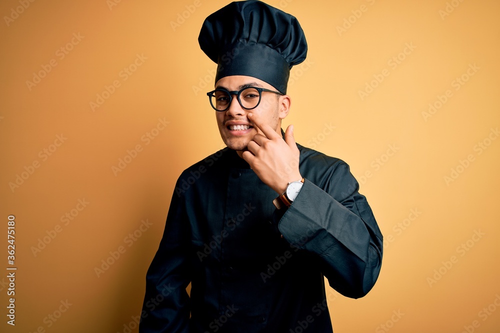 Young brazilian chef man wearing cooker uniform and hat over isolated yellow background Pointing with hand finger to face and nose, smiling cheerful. Beauty concept Young brazilian chef man wearing cooker uniform and hat over isolated yellow background Pointing with hand finger to face and nose, smiling cheerful. Beauty concept