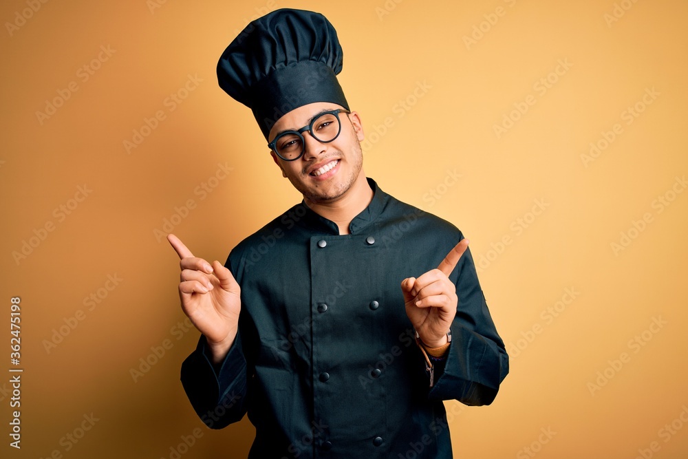 Young brazilian chef man wearing cooker uniform and hat over isolated yellow background smiling confident pointing with fingers to different directions. Copy space for advertisement Young brazilian chef man wearing cooker uniform and hat over isolated yellow background smiling confident pointing with fingers to different directions. Copy space for advertisement