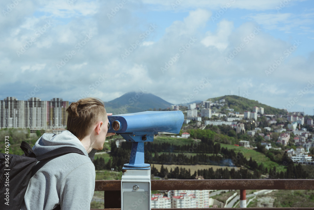 young caucasian hipster man looking through stationary binoculars at sea, mountains and city on observation deck, horizontal outdoors stock photo image