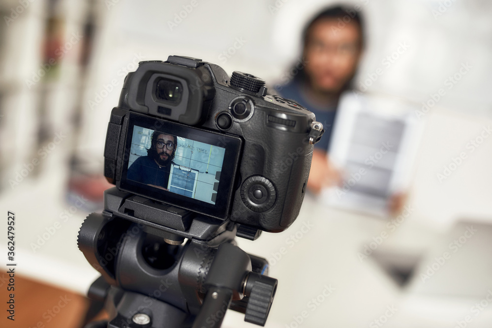 Young man teaching online, looking at camera and explaining new theme while working from home. Focus on camera, professional digital equipment